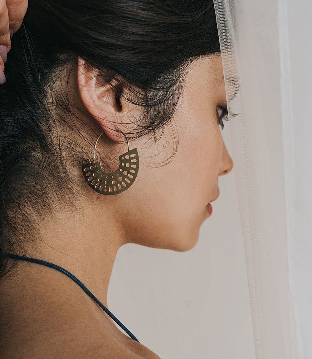 A woman with dark hair shows off a gold hoop earring with artful cutouts as she stands amongst the curtains in her window.