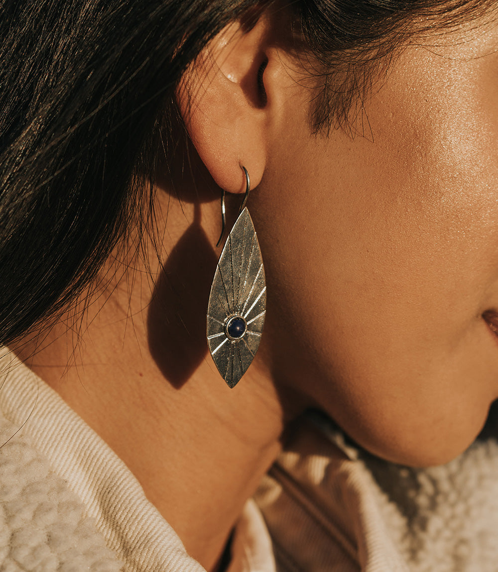 A woman wears silver leaf-shaped earrings with sunburst etchings and blue stone accents.