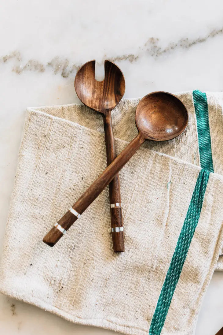 Two dark wood salad servers with silver stripe inlays on the handles rest on a white and blue linen kitchen towel on a marble counter.